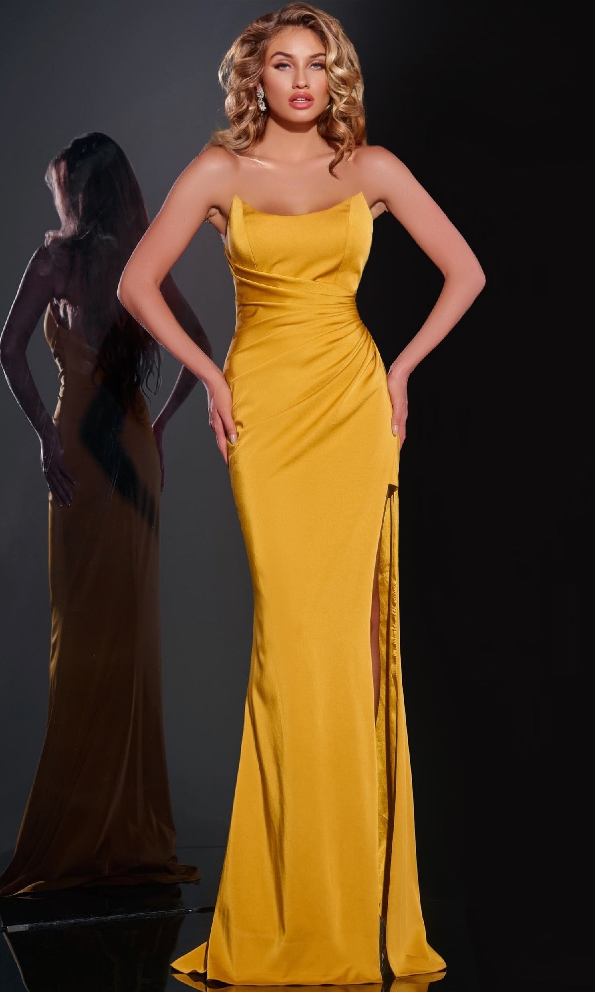 Wearing the Jovani 42765 long prom dress, a woman poses before a black background. The satin, strapless gown features a floor-length silhouette and a thigh-high slit, with her reflection visible behind her.